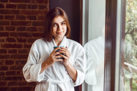 Beautiful caucasian girl, in a white bathrobe, enjoys morning coffee time in the kitchen near windowの写真素材
