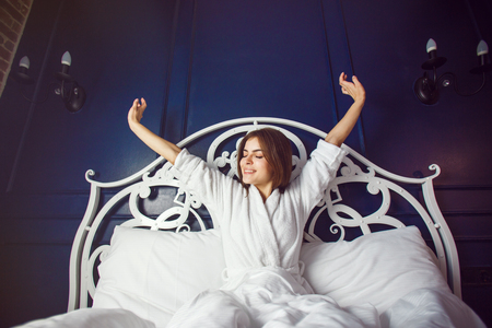 Beautiful caucasian girl, in a white bathrobe, enjoys morning in the bedroom before blue wallの写真素材