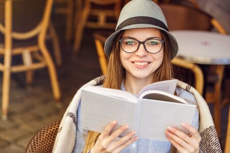 Stylish cheerful girl wears blue shirt, hat and eyeglasses, plaid, reading book in street cafe, outdoors sunny autumn timeの写真素材