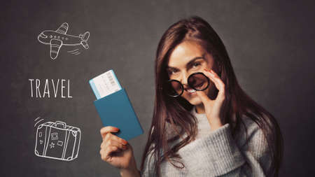 Beautiful long-haired girl wearing sunglasses holding passport with ticket on grey background with travel iconsの写真素材