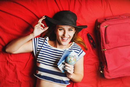 Cheerful girl holding passport, plane ticket and globe lying on red bed with suitcase, indoor travel conceptの写真素材