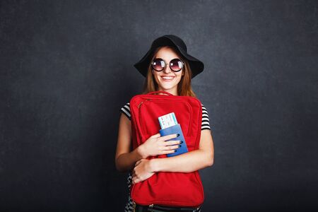 Cheerful happy girl wears black hat and sunglasses holding passport, plane ticket and red suitcase before grey background, indoor travel conceptの写真素材