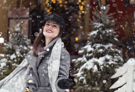 Beautiful stylish girl wears black cap and grey coat having fun with first snow before garland trees, snowy city christmas outdoor shotの写真素材