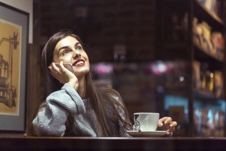 Young pretty brunette girl wears grey sweater have pleasant conversation by smartphone sitting near cafe windowの写真素材