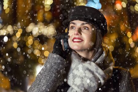 Stylish brunette smiling girl having pleasant talk by smartphone before garland wall in city snowy...