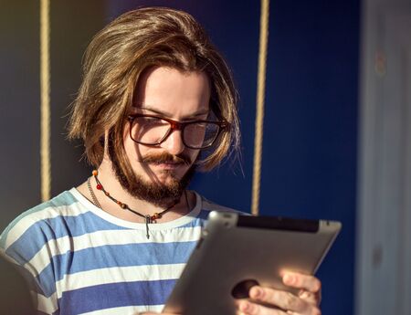 Bearded man in striped t-shirt using tablet in a dark room, indoor shot in the sunlightの写真素材