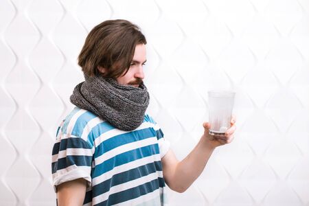 Ill dark-haired man throwing a pill into a glass with water, wearing gray scarf and striped t-shirt,...
