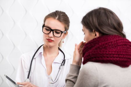 Dark-haired woman in warm scarf and gray top listening to a young medical practitioner, indoor shot in the white backgroundの写真素材