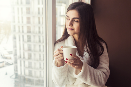 Beautiful girl wears white bathrobe holds cup of coffee sitting on the windowsill near windowの写真素材