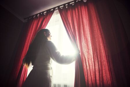 Beautiful girl wears white bathrobe opening curtains on the morning window with sunriseの写真素材