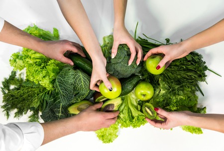 Man and two women picking up fresh green fruit and vegetables from white table, above shot of rich nutritious foodの写真素材