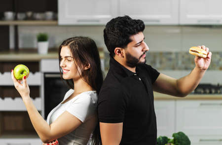 Attractive dark-haired man in black t-shirt and brunette girl hesitating between green apple and tasty hamburger, decides to eat the fresh healthy fruit, concept of choosingの写真素材