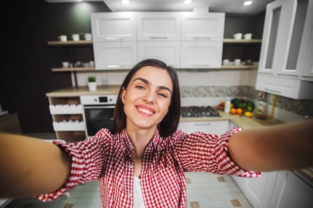 Beautiful brunette in red checked shirt having video talk, smiling girl recording video with a smartphone in light, furnished kitchenの写真素材
