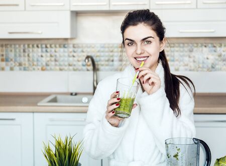 Beautiful brunette enjoying a glass of green smoothie at the kitchen table, concept of healthy lifestyle, peaceful morning timeの写真素材