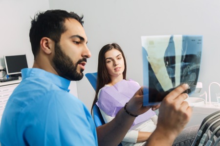 Dentist using x-ray during dental treatment, pointing at the teeth scan and explaining the problem, pretty woman in white top sitting in dental chair at the doctorsの写真素材