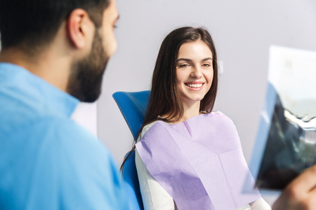 Dentist using x-ray during dental treatment, pointing at the teeth scan and explaining the problem, pretty woman in white top sitting in dental chair at the doctorsの写真素材