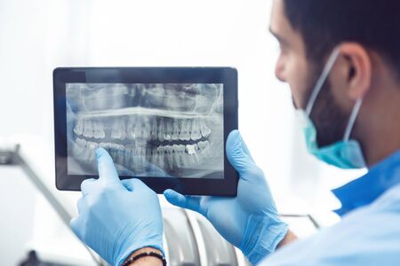 Intelligent dentist showing teeth x-ray on tablet, well-built man in green mask and blue gloves explaining particular case, sitting before dental chair in light huge officeの写真素材