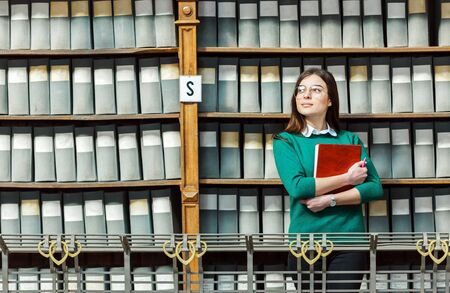 Slender brunette wearing stylish glasses and green sweater holds red book near library bookshelves with grey boxesの写真素材