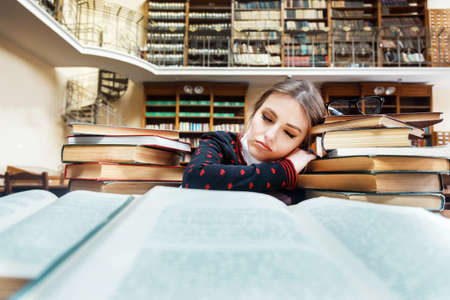 Tired sleeping girl student studying at the library table full of booksの写真素材
