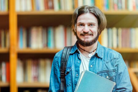 Portrait of young bearded man student holding books before bookshelves in the libraryの写真素材