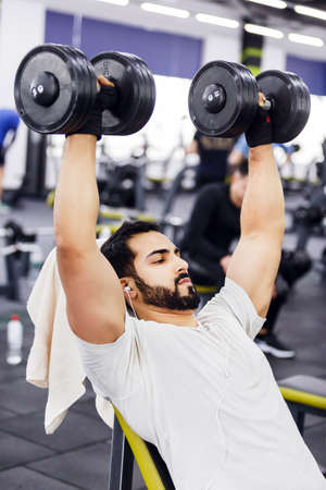 Bearded man wears white t-shirt and headphones does pushup workout with dumbbells on the branch in the gymの写真素材