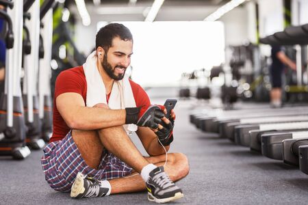 Bearded muscular man wears red t-shirt sitting on floor while rest with towel on neck, using...