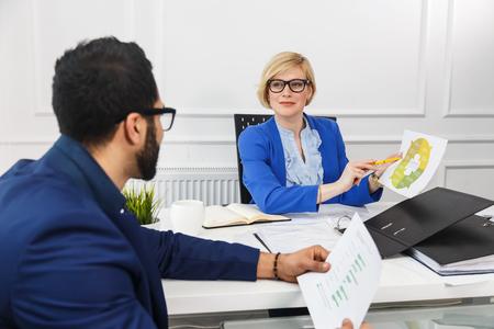 Colleagues working with infographic documents in white office room, bearded man and blonde woman wears eyeglasses, teamwork conceptの写真素材