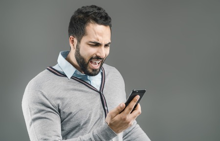 Bearded handsome man with angry face expression holding smartphone in hand on grey isolated...