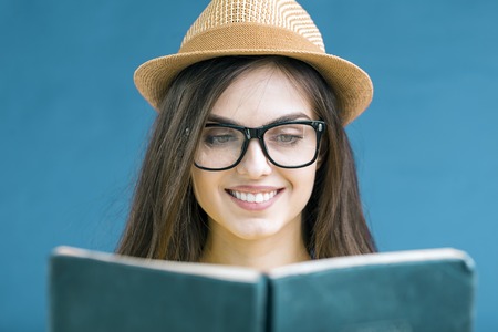 Portrait of beautiful smiling brunette girl wears eyeglasses reading the book with face expression, isolated on blue backgroundの写真素材