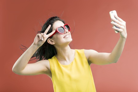 Beautiful smiling brunette girl wears yellow t-shirt and sunglasses making selfie by smartphone before red backgroundの写真素材