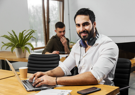 Attractive bearded man in pristine white shirt working on silver laptop, wearing nice big headphones around the neck, two other empoyees doing the task behind the manの写真素材