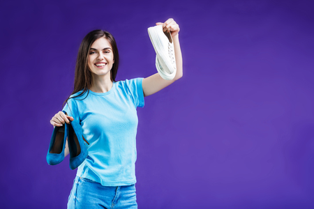 Beautiful brunette girl dressed in blue t-shirt choosing between comfortable sneakers shoes and heels before blue background, smart choise conceptの写真素材
