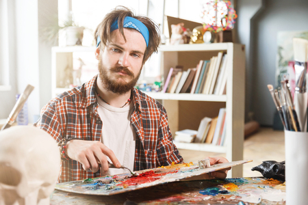 Portrait of a young bearded male artist is holding a brush and mix color oil painting on paletteの写真素材
