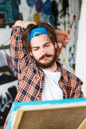 Handsome dark-haired bearded hipster male artist in blue bandana sitting and looking on canvas in his light modern studioの写真素材