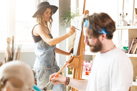 Caucasian dark-haired hipster male artist in blue bandana sitting and drawing with brush on canvas in his light modern studio, his colleague drawing behind the easel near the windowの写真素材