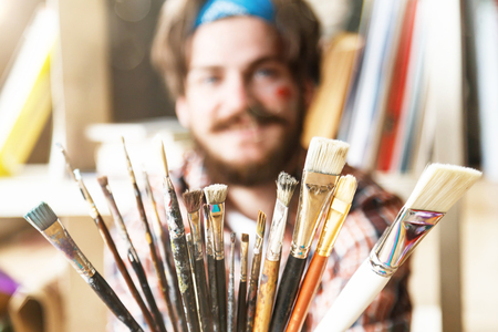 Dark-haired caucasian bearded hipster male artist in blue bandana hoilding many brushes in light studioの写真素材