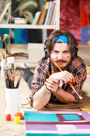 Caucasian dark-haired bearded hipster male artist in headphones and blue bandana lies on the floor and holding brush in his contemporary studio with many paintingsの写真素材