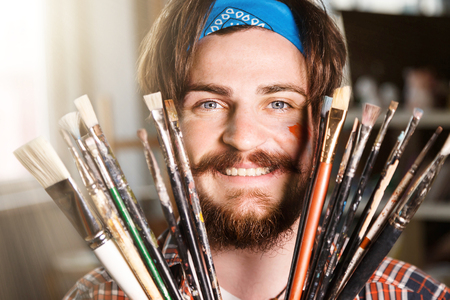 Dark-haired caucasian bearded hipster male artist in blue bandana hoilding many brushes in light studioの写真素材