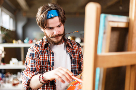 Creative dark-haired bearded hipster male artist in blue bandana holding palette and drawing behind the easel in his contemporary studio with many paintings, holding a brush in his teethの写真素材