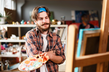 Dark-haired bearded hipster male artist in blue bandana holding brush in his teeth and looking on the canvas behind the easel in his contemporary studio with many paintings,の写真素材
