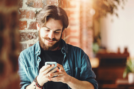 Handsome young bearded boy reading sms on the mobile phone and smiling near the window in the loft modern officeの写真素材