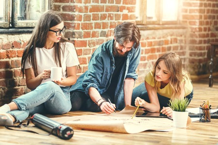 Crew of young professional architects discussing blueprint of new modern house on the floor in the loft light officeの写真素材