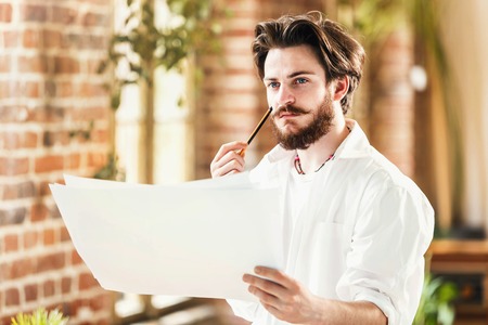 Bearded male architect in white shirt thinking and analyzing the details of his project in loft...