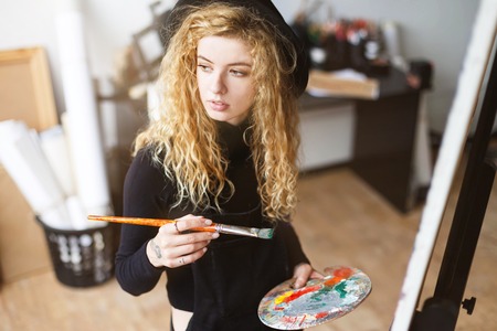 Blonde curly feemale artist in black hat holding brush and palette with oil behind the easel in light studio, she looking to the windowの写真素材