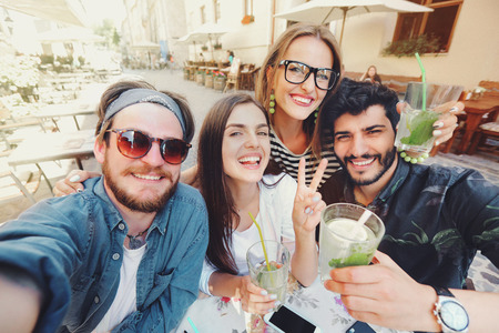 Four joyful cheerful friends taking a selfie while sitting together at cafe and showing peace gesture outdoorsの写真素材
