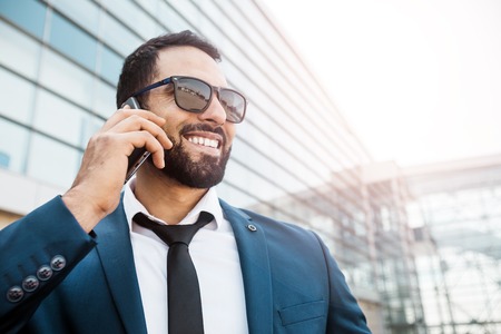 Bearded smiling businessman wears blue suit and sunglasses talking via smartphone before high modern buildingの写真素材