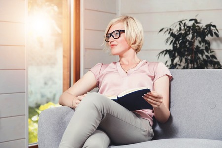 Pretty blonde woman in glasses sitting with book on the gray sofa near the fireplace in the comfortable living-room and looking on the windowの写真素材