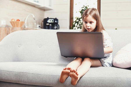Pretty little brunette girl with excited face expression sitting on the sofa and using Internet on the laptopの写真素材