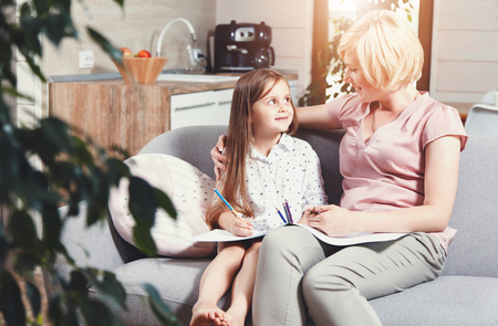 Pretty blonde mother and cute brunette daughter sitting on the sofa and drawing pictures with color pencilsの写真素材
