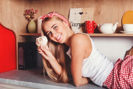 Pretty blonde long hair woman laying on the table, smiling and holding cupcake in the red loft comfortable kitchenの写真素材